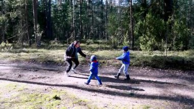 İki çocuk sevinçle orman yolunda koşarken bir ebeveyn yakınlarda çömelmiş ağaçlarla çevrili, ferahlatıcı açık havada hayat dolu bir aileyi yakalıyor..