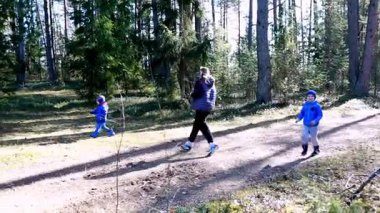 Bir anne ve iki çocuğu, uzun ağaçlar ve parlak güneş ışığıyla çevrili bir orman yolunda neşeyle koşuyorlar. Neşeli atmosfer onların mutlu etkileşimlerini vurgular..