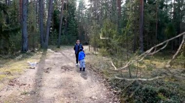 Bir anne ve oğlu ormanlık bir yolda geziniyor, doğanın tadını çıkarırken küçük bir köpek de yakınlarda geziniyor. Güneş ağaçların arasından süzülür ve sakin bir atmosfer yaratır..