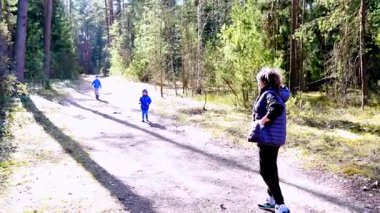 İki çocuk sakin bir orman yolunu keşfederken bir yetişkin uzaktan izliyor. Güneş ağaçların arasından parlıyor, sıcak ve davetkar bir atmosfer yaratıyor..