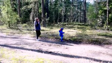 Bir anne ve küçük çocuğu, uzun ağaçlar ve canlı yeşilliklerle çevrili bir orman patikası boyunca yürürler. Güneş parlıyor, sakin bir atmosfer yaratıyor. Çocuk mutlu bir şekilde çevreyi keşfediyor..