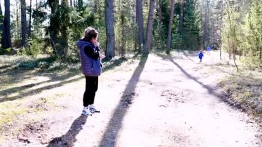 Bir kadın ağaçlık bir yolda duruyor, çocuklar uzaklarda oynarken sohbete dalıyor. Güneş dalların arasından ışıl ışıl parlıyor, canlı bir atmosfer yaratıyor..