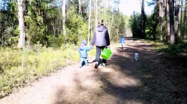 Bir kadın ve küçük bir çocuk yeşil bir kova taşıyarak orman yolunda el ele yürürken, diğer iki çocuk da yakınlarda oyun oynuyor. Küçük bir köpek sevinçle arkadan geliyor..