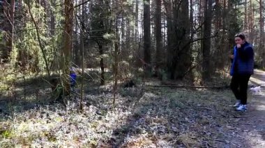 Bir çocuk ağaçların arasında mutlu bir şekilde oynarken bir yetişkin yanında duruyor ve ormanın huzurlu ortamını seyrediyor. Güneş ışığı yaprakların arasından süzülür ve dışarıda güzel bir günün tadını çıkarırlar..