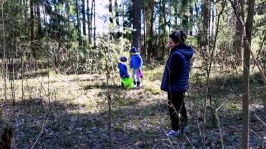 Sıcak giyimli iki çocuk doğal hazineler için ormanı ararken bir yetişkin yakınlarda gözcülük yapıyor. Güneş ışığı ağaçların arasından süzülüp, barışçıl bir atmosfer yaratır..