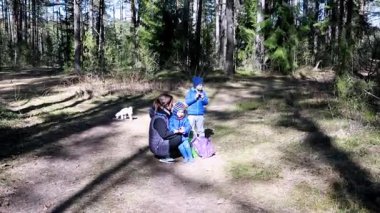 Bir anne, dışarıda ormanda oynarken çocuklarına yardım eder. Aydınlık bir gün ve bölge yüksek ağaçlar ve yeşilliklerle çevrili. Yakında küçük bir köpek var..