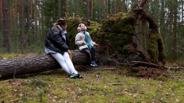 Çocuklar canlı yeşil bir ormandaki büyük bir devrilmiş ağaçta otururken huzurlu bir anın tadını çıkarırlar. Sıkı giyinmişler, doğa ve sonbahar renkleriyle çevrelenmişler..