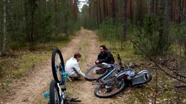 Sakin bir ormanda, iki bisikletçi ağaçlarla kaplı pis bir patikada bisikletlerini tamir etmek için birlikte çalışırlar. Atmosfer sakin ve odaklanmış, doğada takım çalışması sergiliyor..