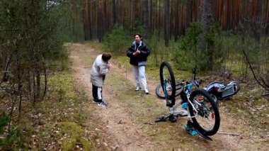 İki arkadaş, çamurlu bir patika boyunca bisiklet sürdükten sonra sakin bir ormanda biraz dinlenir. Yeşillik ve uzun ağaçlar doğanın tadını çıkarmak için mükemmel bir ortam yaratır..