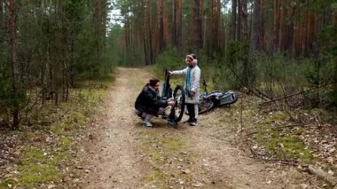 İki bisikletçi tenha bir orman yolunda bisiklet lastiğini tamir etmek için birlikte çalışır. Etraflarını saran uzun ağaçlar açık hava maceraları için sakin bir atmosfer yaratıyor..