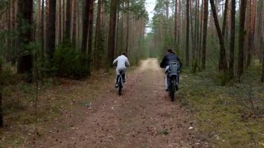 İki birey, uzun ağaçlar ve serin bir bahar gününde doğanın sesleriyle çevrili sakin bir orman ortamında ağaçlık bir patika boyunca bisiklet sürüyor..