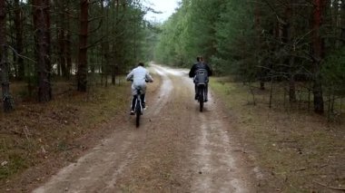 İki birey, uzun ağaçlarla çevrili huzurlu toprak patika boyunca bisiklet sürüyor. Güneş yaprakların arasından parlıyor, açık hava macerası için sakin bir atmosfer yaratıyor..