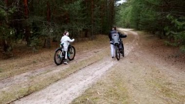İki birey, uzun ağaçlarla çevrili yemyeşil bir ormandaki kirli bir yolda bisiklet macerasından zevk alıyor. Atmosfer huzurlu, aile dostu bir açık hava aktivitesi sergiliyor..
