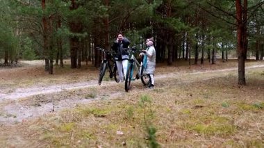 İki kişi sakin bir ormanda bisikletleriyle ayakta duruyor. Manzara, bereketli ağaçlar ve sakin bir gezinin özünü yansıtan yumuşak, topraksı bir yol içerir..