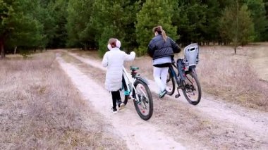 İki birey, uzun ağaçlarla çevrili toprak bir patika boyunca sakin bir bisiklet yolculuğunun tadını çıkarıyor. Bir kişi bisiklete binerken diğeriyle sohbet eder..