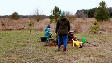 Bir çocuk, seyrek ağaçlar ve çimlerle çevrili açık bir arazide bakıcıya ağaç dikmesine yardım eder. Bulutlu gökyüzü bu ilkbahar aktivitesine sakin bir atmosfer ekler..