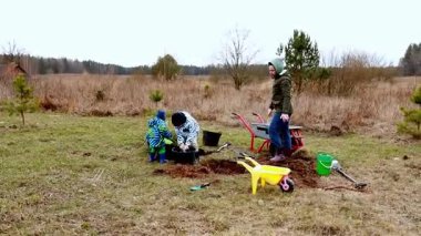 Renkli ceketli çocuklar bir yetişkinin açık arazide çiçek açmasına yardım ediyor. Takım çalışmasını ve doğa takdirini teşvik eden eğlenceli bir doğa aktivitesiyle uğraşırken toprak ve araç topluyorlar..