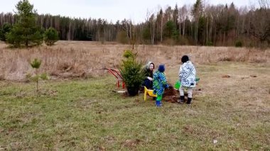 Kışın giyinmiş iki çocuk çimenli bir tarlaya küçük bir ağaç dikiyorlar. Manzara, doğanın güzelliğini gözler önüne seren seyrek ağaçlara ve açık alana sahiptir..