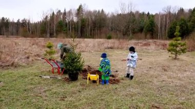 İki küçük çocuk ve bir yetişkin çimenli bir alanda birlikte ağaç dikmeye katılıyorlar. Serin bahar havası aile bağlarını kuvvetlendiriyor..