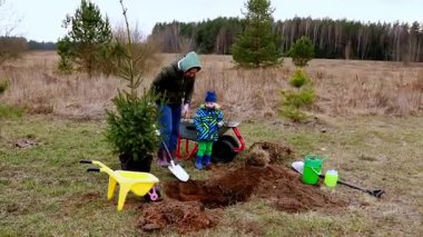 Bir kadın ve bir çocuk banliyö tarlasına ağaç dikmek için birlikte çalışıyorlar. Hoş bir bahar gününün tadını çıkarırken bahçe aletlerini kullanıyorlar. Çevrede yeşillik ve ağaçlar var..