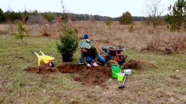 Bir kadın, ağaçlarla çevrili sakin bir açık alana genç bir ağaç dikmek için çukur kazıyor. Yakınlarda bir çocuk izliyor, ve bahçe aletleri etrafa saçılmış, canlı bir açık hava aktivitesi sergiliyor..