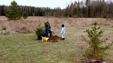 Bir çocuk ve bir yetişkin ağaçlarla çevrili çimenli bir alana ağaç dikiyorlar. Soğuk hava için sarılıp sarmalanmışlar. Açık hava aktivitelerinin tadını çıkarırken olumlu bir etki yaratmışlar..