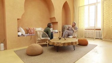 Sıcak tonların ve doğal malzemelerin yer aldığı sıcak bir odada, iki kadın rahat bir şekilde otururken anlamlı bir sohbete girişiyor. Rahatlama atmosferi onların etkileşimini güçlendirir..