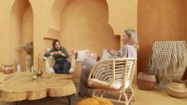 İki kadın, sıcak renkler ve doğal malzemelerle süslenmiş, kapalı bir alanda iç içe geçmiş bir şekilde sohbet ediyorlar. Atmosfer davetkar ve rahat, hikayeleri paylaşmak için mükemmel..