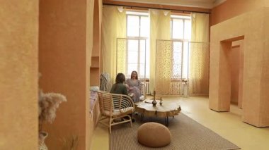 Ilık tonlu ve doğal ışıkla donatılmış bir ortamda iki kişi sakin bir tartışma içindeler. Mekanda rahat mobilyalar ve barışçıl bir atmosfer yaratan dekoratif elementler bulunuyor..