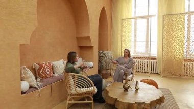 Sıcak tonlarla süslenmiş karşılama alanında iki kadın, şık sandalyelerde rahat bir şekilde otururken ve doğal dekor elementleriyle çevrelenmişken derin bir sohbetten zevk alırlar..