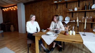 Sıcak, davetkar bir odada, iki kadın yemek takımları ve mumlarla süslenmiş ahşap bir masada otururken ev dekorasyonu hakkında sohbet ediyorlar..
