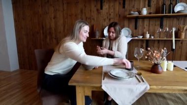 Sıcak bir restoran atmosferinde, iki kadın yemek yerken hareketli bir sohbete girişir, kahkahaları paylaşır ve şık ahşap bir masada birbirlerine eşlik ederler..