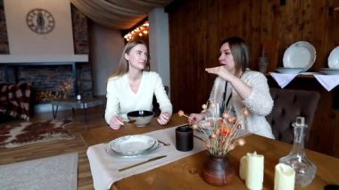 İki arkadaş şık yemek takımlarıyla dolu bir masada oturup sıcak dekorasyonlarla süslenmiş, sıcacık, kırsal bir ortamda hafif bir yemek yerken hikayeler ve kahkahalar atıyorlar..