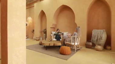 Sıcak ve davetkar bir odada, toprak tonlarıyla ve doğal malzemelerle süslenmiş iki kadın, dostça bir sohbete girişiyor. Dekorasyonda desenli duvarlar ve şık dekorlar yer alıyor.