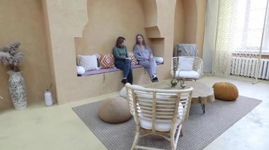 İki kadın, doğal malzemeler ve sıcak renklerle dizayn edilmiş sakinleştirici bir salonda oturuyorlar. Sağlıklı yaşam çekilmesinin huzurlu atmosferinin tadını çıkarırken sohbet ediyor gibi görünüyorlar..