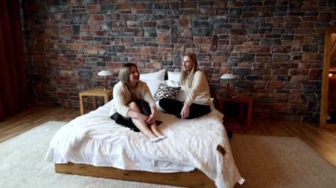 İki arkadaş şık bir odadaki bir yatakta rahat bir şekilde oturuyorlar. İçinde tuğla duvarlar ve modern mobilyalar var. Gülüyorlar, sohbet ediyorlar. Birlikte rahat bir ortam yaratıyorlar..