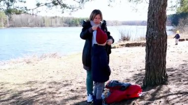 Bir anne, küçük oğluna göl kenarında yardım eder. Giysilerini açık hava aktivitelerine hazırlanırken ayarlar. Güneş parlıyor ve doğa onları kuşatıyor..