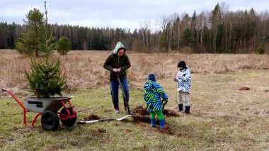 Bir kadın ve iki çocuk serin bir bahar günü ağaçlarla çevrili güzel bir açık alanda ağaç dikme etkinliğine katılıyorlar..