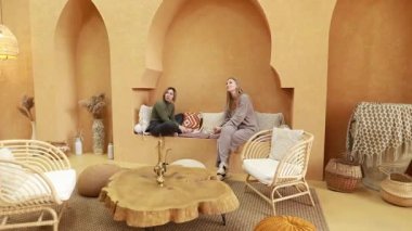 İki arkadaş, modern mobilya ve doğal aksanlarla dolu sıcak tonlu bir odada sakin ve davetkar bir atmosfer yaratarak keyifli bir sohbetin tadını çıkarıyorlar..