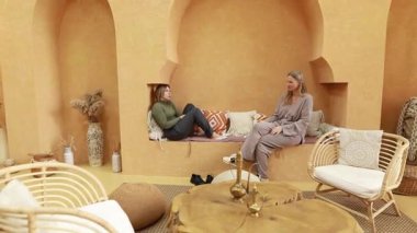 İki kadın güzel tasarlanmış bir odada rahat bir şekilde otururlar. Sıcak tonlarda ve rahat bir dekorasyonla..