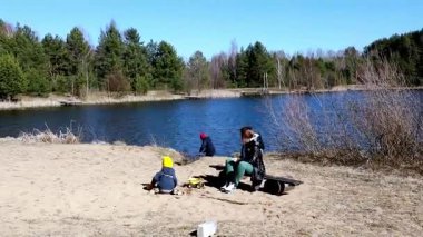 Bir aile göl kenarında güneşli bir öğleden sonra geçirir, çocuklar kumsalda oynar ve dışarıda eğlenir. Kahkaha ve doğa neşeli bir atmosfer yaratır..