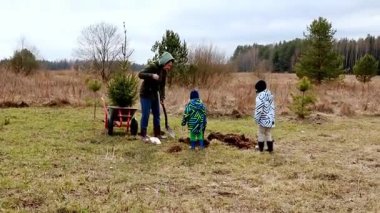 Bir aile ağaç dikmekle meşgul olurken, iki çocuk bu süreçte yardımcı oluyor ve eğleniyor. Ağaçlarla çevrili çimenli bir alandalar, çevresel bakımı destekliyorlar..