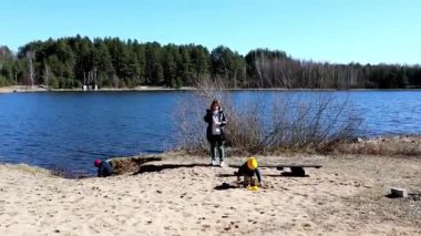 İki genç çocuk dingin bir gölün kenarında eğlenirken yetişkinler neşeli anlarını kaydediyor. Açık gökyüzü ve çevre doğa aile eğlencesi için sakin bir atmosfer yaratıyor..