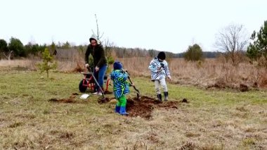 İki çocuk çimenli bir alana genç bir ağaç dikmeye yardım ederken bir aile dışarıda vakit geçirir. Renkli ceketler giyiyorlar, serin havada takım çalışması ve çevresel bakım yapıyorlar..
