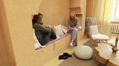 İki kadın, rahat bir yaşam alanında rahat bir şekilde, doğal ışık ve sıcacık renklerle dolu, rahat mobilyalar ve düşünceli dekorlarla çevrili hayattan bahsederek rahat bir atmosfer yaratıyor..