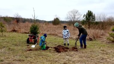 Bir grup çocuk kırsal bir alana hevesle genç ağaçlar dikerler. Bulutlu bir günde açık havada eğlenirken çukur kazıp zemin hazırlarlar..