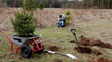 Sakin bir tarlada, bir aile anın tadını çıkarırken genç bir ağaç diker. Bir çocuk ve yetişkin, yeni kazılmış toprak ve bahçe aletlerinin yanında samimi bir kucaklaşmayı paylaşıyor..