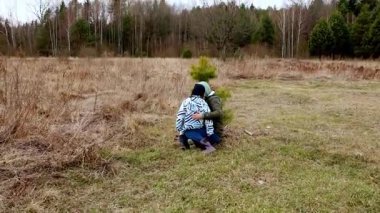 Güneşli bir tarlada oynayan iki çocuk, küçük yeşil bir ağacı kucaklıyor. Çevrede uzun otlar ve seyrek ağaçlar bulunur, sakin bir açık hava ortamı oluşturur..