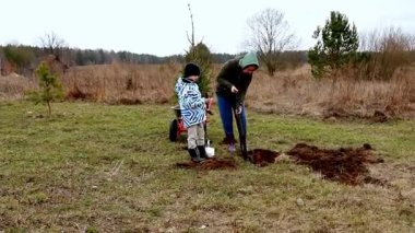 Bir kadın ve bir çocuk çim tarlasına ağaç dikmek için toprağı kazarak bahçıvanlık yapıyorlar. Serin bir gün için sıcacık giyinirler ve arka planda seyrek ağaçlar bulunur..