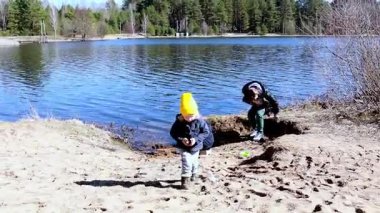 İki çocuk kumsalda neşeyle oynarken bir yetişkin yakınlarda bir aktiviteye girişiyor. Güzel göl parlak güneş ışığı altında ağaçlarla çevrilidir..